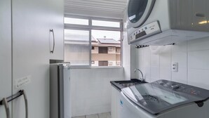 A compact laundry room with a washing machine, dryer, and a window providing a view of the outside. - Apt W/bbq and Access to Praia Brava Rra0103 (Florianópolis)
