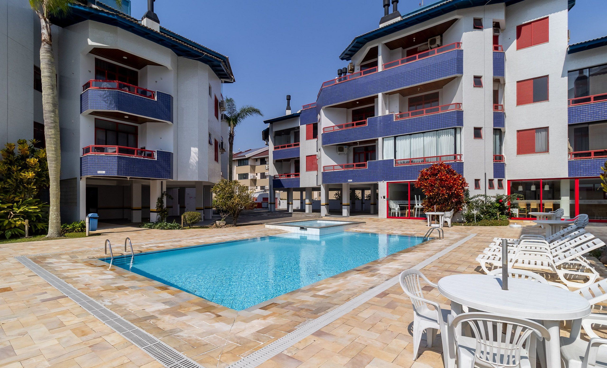 A swimming pool area with tiled flooring, lounge chairs, and a building with balconies.