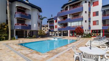 A swimming pool area with tiled flooring, lounge chairs, and a building with balconies.