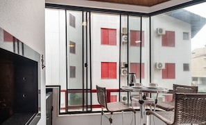 A balcony with a table and chairs, a view of a building with red shutters, and an air conditioning unit.
