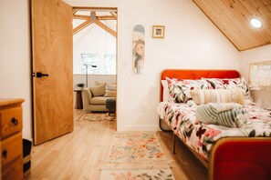 Un dormitorio con puerta de madera, una cama con ropa de cama floral, cabecera roja y vista a otra habitación con un sofá y una ventana.