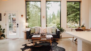 A modern living room with a black leather sofa, a wooden coffee table, and a cowhide rug. Large windows offer views of a forested area.