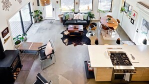 A spacious living area with a high ceiling, wooden beams, large windows, a fireplace, and a kitchen with a gas stove.