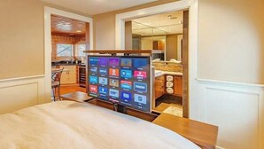 Une chambre d’hôtel avec un lit, un téléviseur affichant les logos des chaînes, une kitchenette et une salle de bain visible à travers une porte ouverte.
