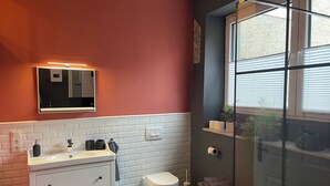Une salle de bain moderne avec un lavabo, un miroir et des toilettes blancs, une douche en verre et une fenêtre avec des stores.