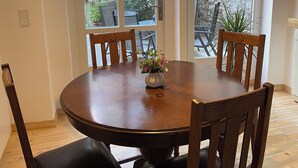 Un espace repas comprenant une table ronde en bois, six chaises en bois, un luminaire suspendu et une vue sur une terrasse extérieure, accessible par de grandes fenêtres en arc de cercle, avec un mur en briques et des plantes en pot.