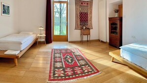 Una habitación con suelo de madera, una cama, una silla, una mesita, un tapiz y un ventanal con cortinas.