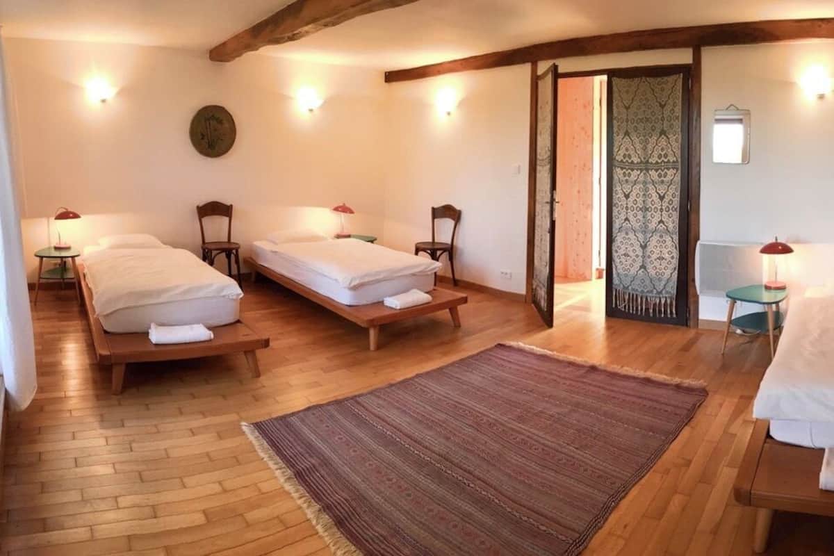A room with three single beds, wooden flooring, and a large rug. There are bedside tables with lamps and a wall-mounted clock.