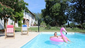 Una piscina con una persona en un flotador con forma de flamenco rosa, rodeada de sillas de descanso y una casa al fondo.