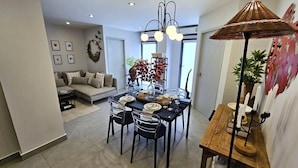 A dining area with a table set for four, surrounded by chairs. There is a sofa in the background and a decorative wall piece above the table. - C212 NOON Village (Chalong)