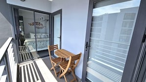 A balcony with a wooden table and chairs, a sliding glass door leading to a bedroom with a bed and a decorative wall piece. - C212 NOON Village (Chalong)