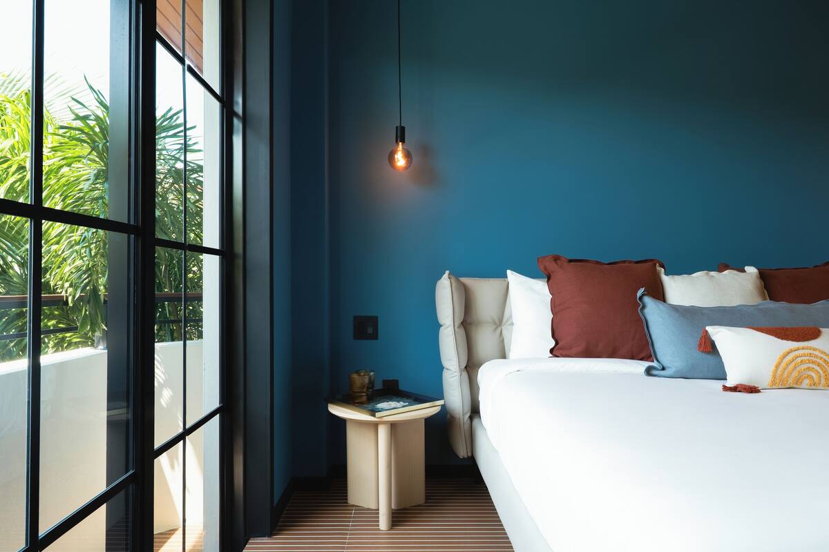 A modern bedroom with a large bed, a bedside table, a hanging light, and a view of greenery outside.