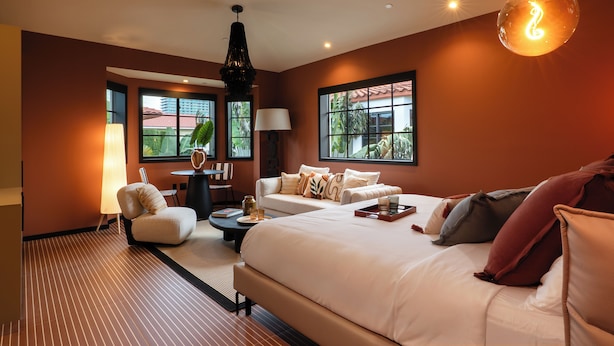 A modern hotel room with a large bed, a sofa, a desk, and a lamp. There are windows with a view of palm trees and a building.
