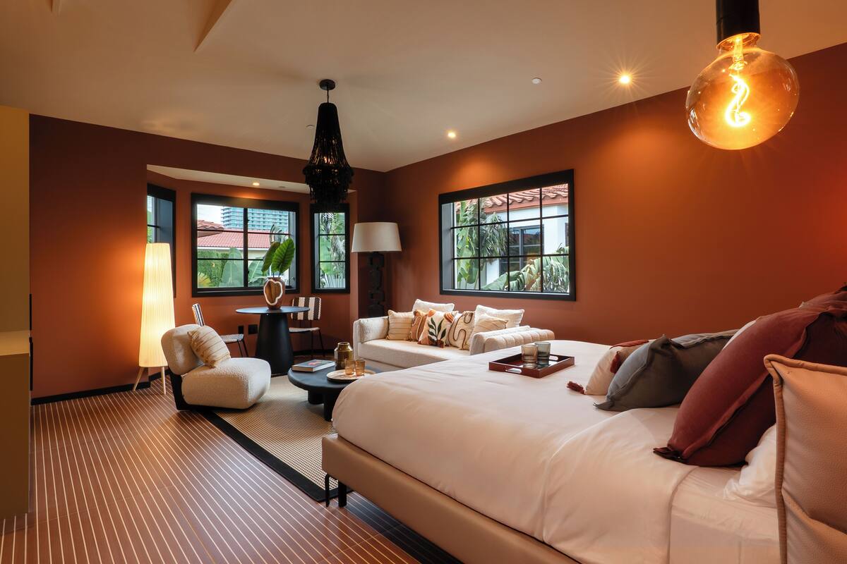 A modern hotel room with a large bed, a sofa, a desk, and a lamp. There are windows with a view of palm trees and a building.