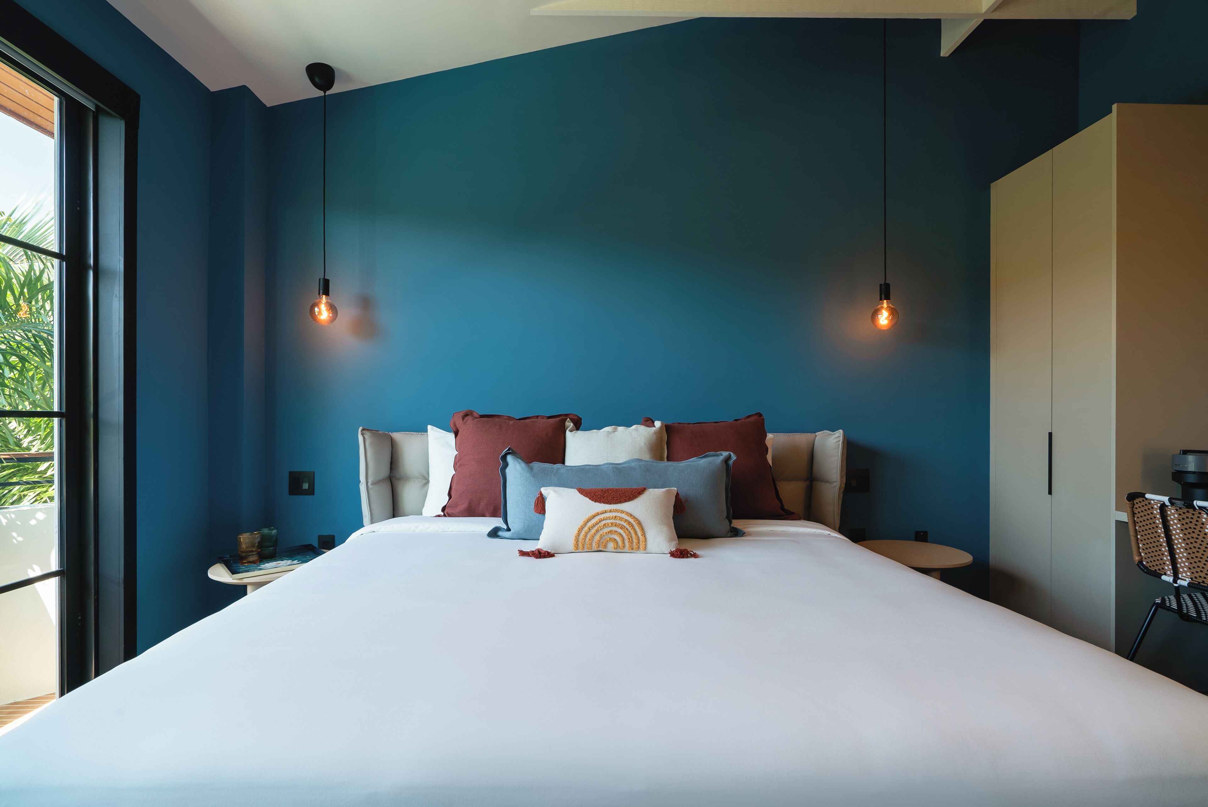 Habitación de hotel moderna con una cama grande, dos lámparas colgantes y vistas a la vegetación por la ventana.