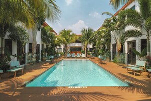 Piscina con sillas de descanso y palmeras, rodeada de edificios blancos con tejados rojos.
