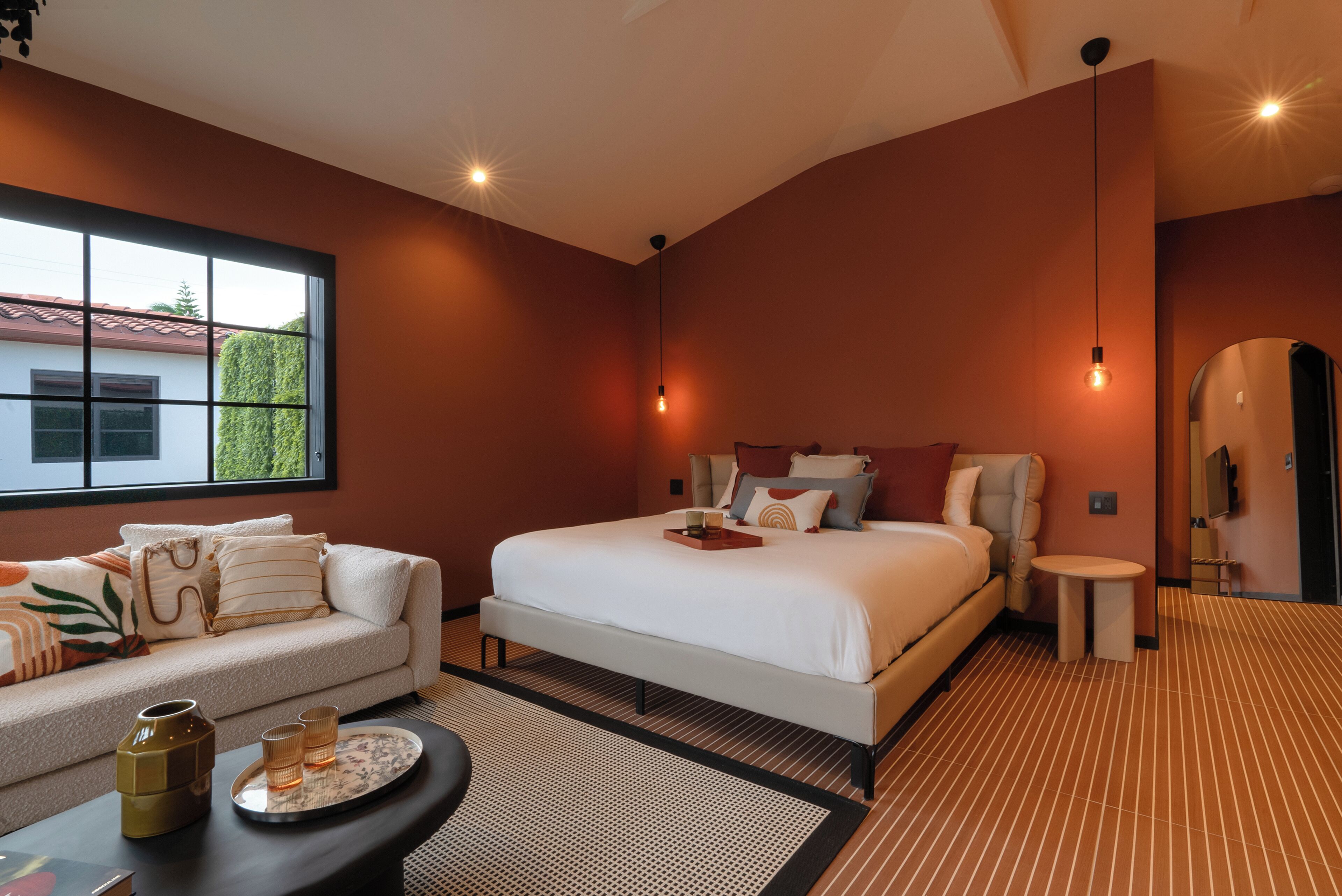A modern hotel room with a large bed, a sofa, a small table, and a large window.