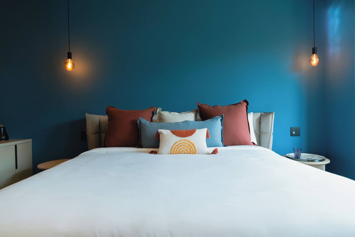 A neatly made bed with multiple pillows, a bedside table with a glass, and two hanging light bulbs.