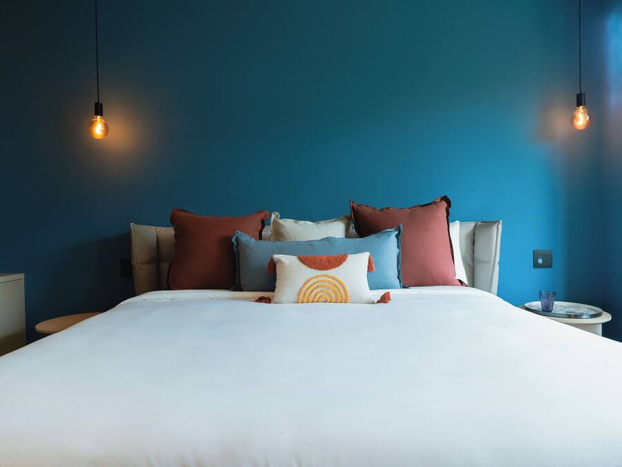 A neatly made bed with multiple pillows, a bedside table with a glass, and two hanging light bulbs.