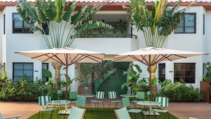 An outdoor patio area with green and white checkered chairs and tables, surrounded by lush tropical plants and a staircase leading to an upper level.