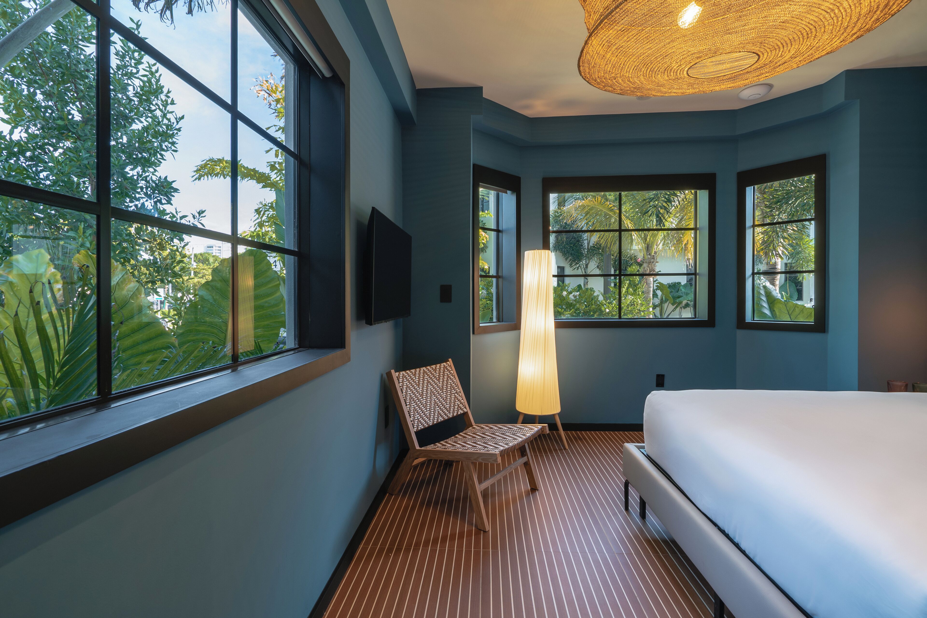 Una habitación con cama, una silla, televisor y ventanas amplias con vistas a zonas verdes.