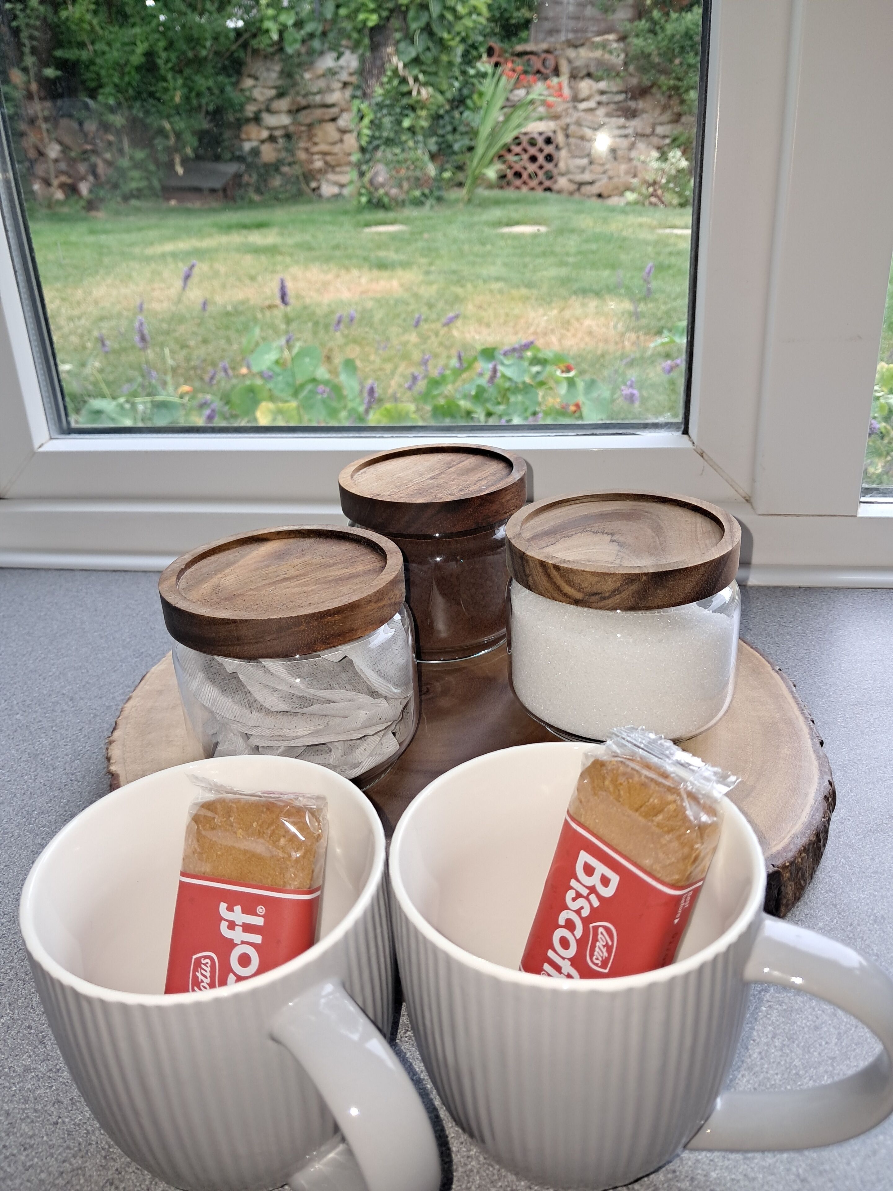 Vier witte mokken met een rood logo, drie houten bakken met deksels, en een glazen pot met een witte deksel op een houten onderzetter.