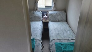 A narrow room with two beds, a small table with a tissue box, and a window with curtains.