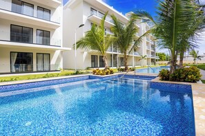 A swimming pool with clear blue water, surrounded by a white building with balconies and palm trees.