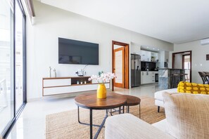 Uma sala de estar moderna com uma televisão de tela plana, um espaço de jantar com almofadas amarelas e uma cozinha ao fundo.