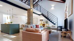 Un salón espacioso con techo alto, una escalera, un mueble verde, un sofá beige y un televisor sobre un soporte de madera.