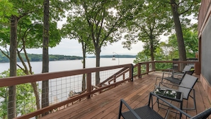 Une terrasse en bois dotée d’une rambarde, qui offre une vue sur un lac où l’on peut apercevoir des bateaux et des arbres.