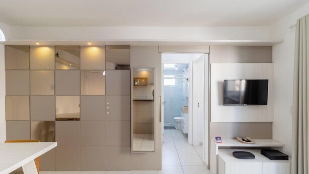 A modern hotel room with a large mirror, a flat-screen TV, a small table with a remote, and a built-in oven.