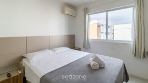 A hotel room with a bed, a bedside table, an air conditioning unit, and a window with curtains.