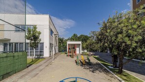 A playground with swings and a slide, a building with a white facade, and a tree-lined street. - Apt. w/ Barbecue & Pool in Canasvieiras Rpc0305 (Florianópolis)