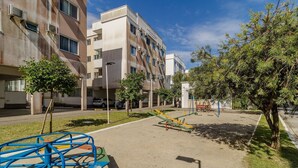 A playground with swings and a slide, surrounded by trees and a building with multiple windows and air conditioning units. - Apt. w/ Barbecue & Pool in Canasvieiras Rpc0305 (Florianópolis)