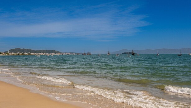 Una playa con aguas tranquilas, barcos y colinas a lo lejos.