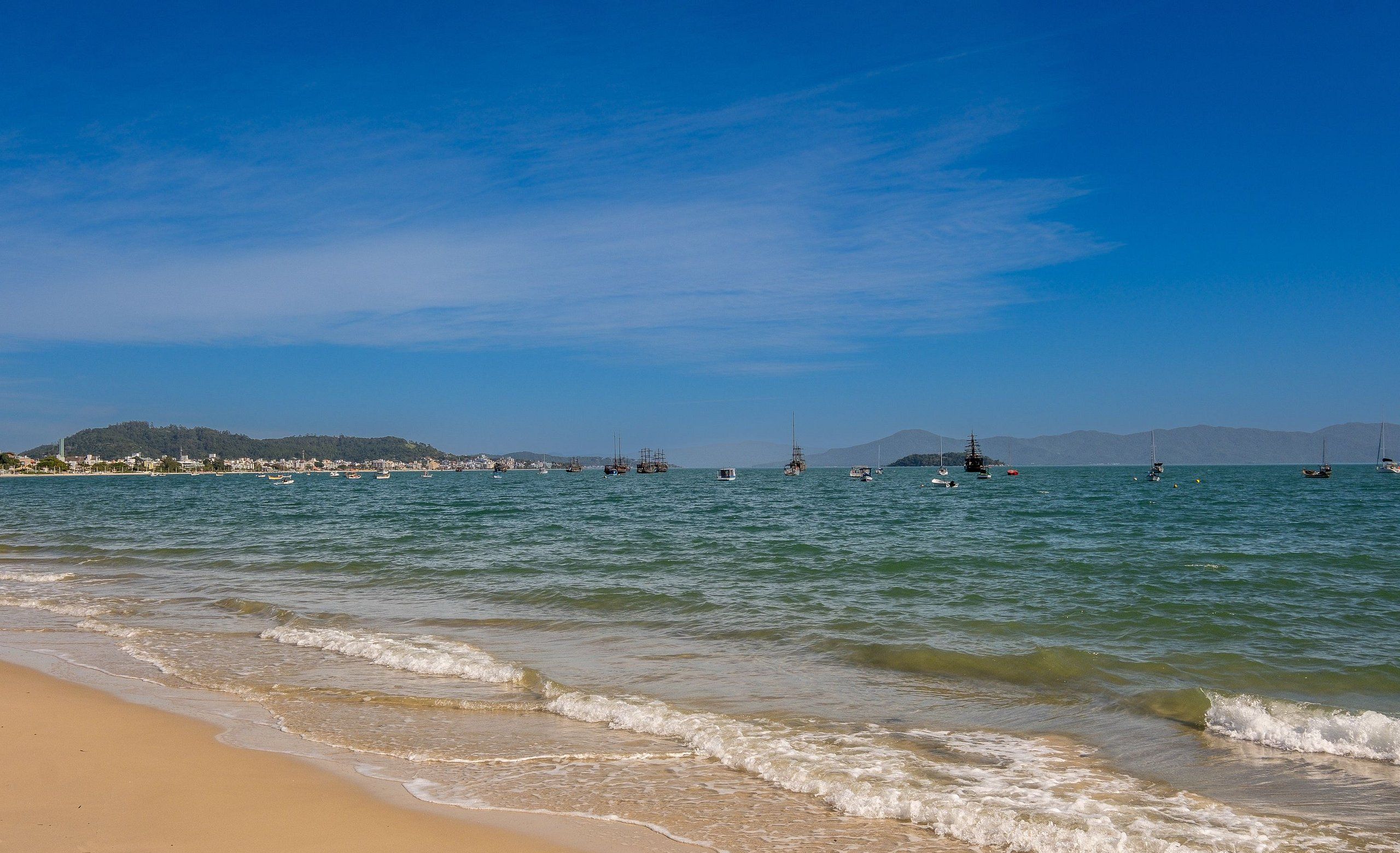 A beach with calm waters, boats, and distant hills.