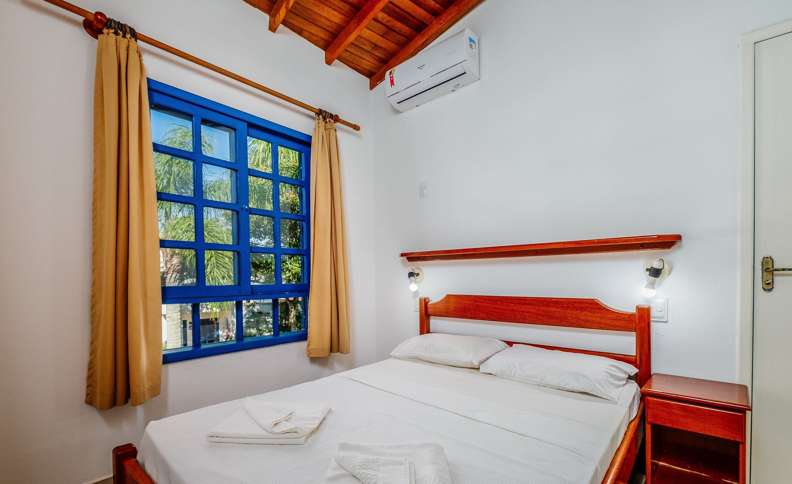 A bedroom with a wooden bed, white bedding, a window with blue frames, beige curtains, and a small bedside table.