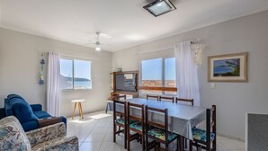 Un comedor con una mesa y sillas, un televisor sobre un mueble, un ventilador de techo y vista al mar a través de amplios ventanales.