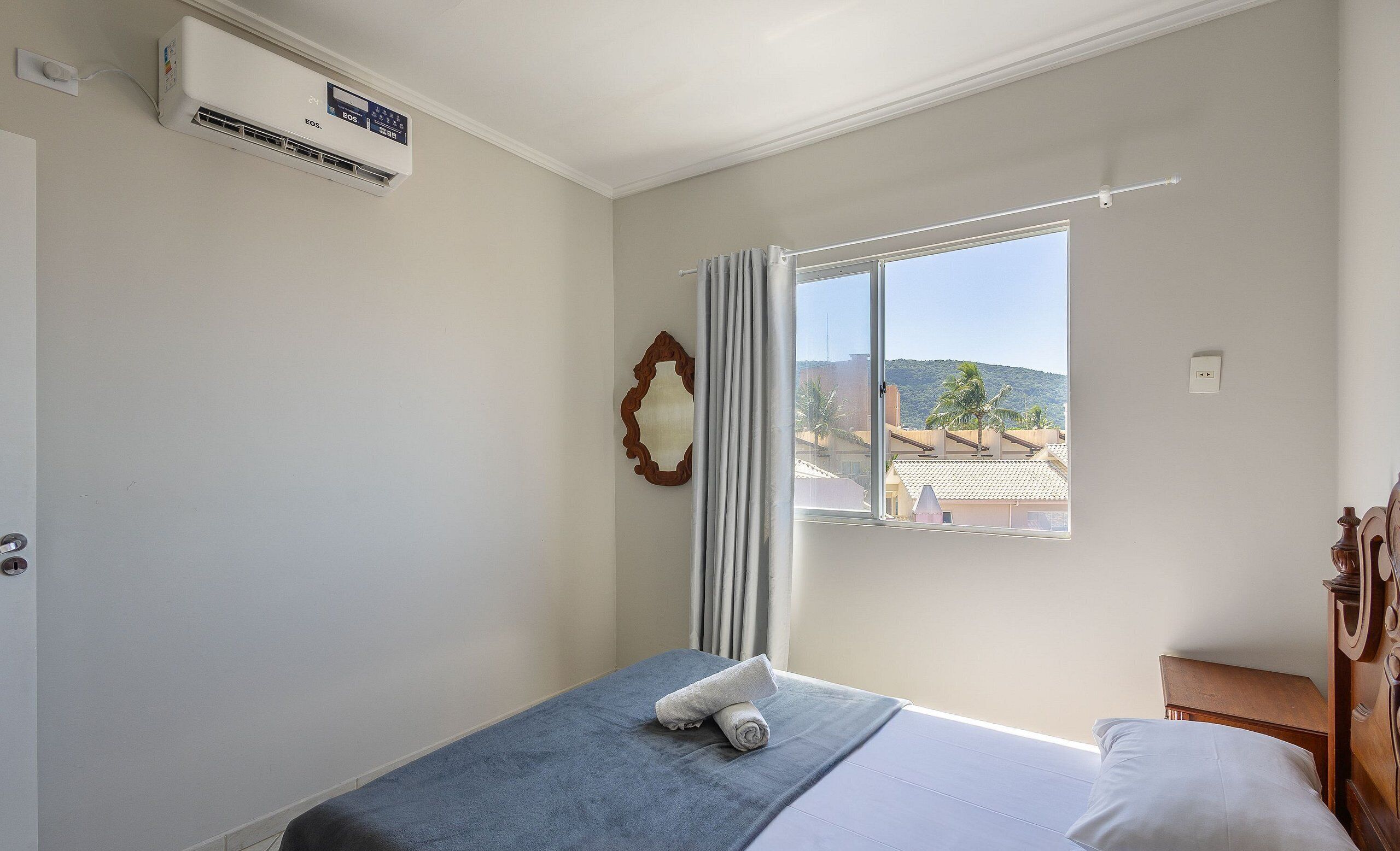 A hotel room with a bed, a window with curtains, an air conditioning unit, and a view of buildings and hills outside.