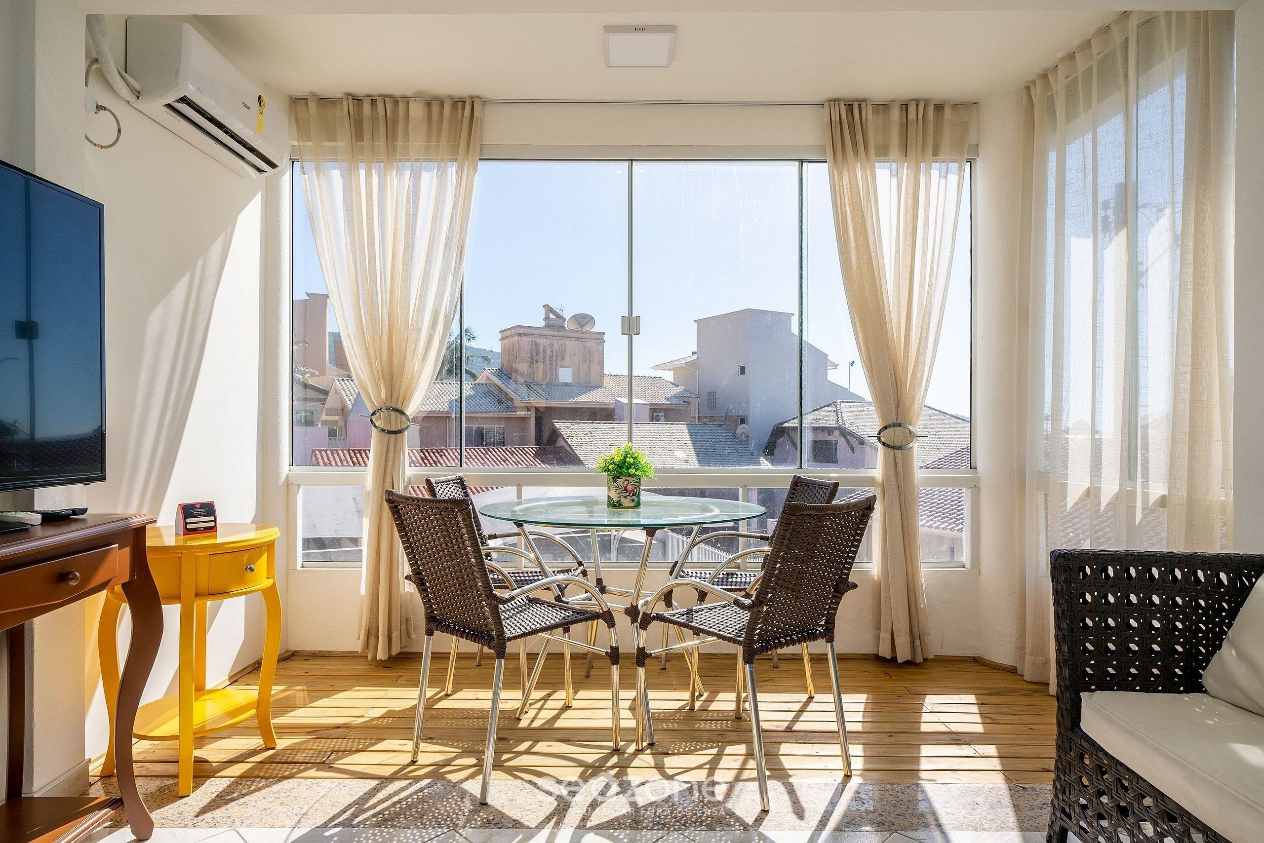 A room with a round glass table, four wicker chairs, a yellow side table, a television, and a window with curtains.