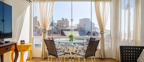 A room with a round glass table, four wicker chairs, a yellow side table, a television, and a window with curtains.