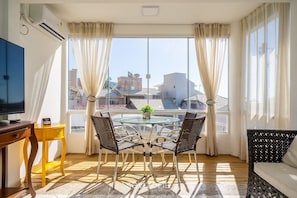 A room with a round glass table, four wicker chairs, a yellow side table, a television, and a window with curtains.