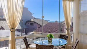 A balcony with a glass table and wicker chairs, overlooking a coastal town with red-tiled roofs. - 2BR apt W/suite 100m Bombas Beach Ror0001 (Bombinhas)