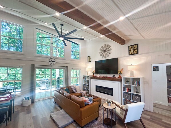 A spacious living room with a large window, a fireplace, a television, and a ceiling fan.