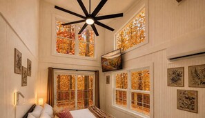 Une chambre d’hôtel avec un lit, un ventilateur de plafond, de nombreuses fenêtres donnant sur des arbres aux feuilles d’automne et des œuvres d’art encadrées accrochées aux murs.