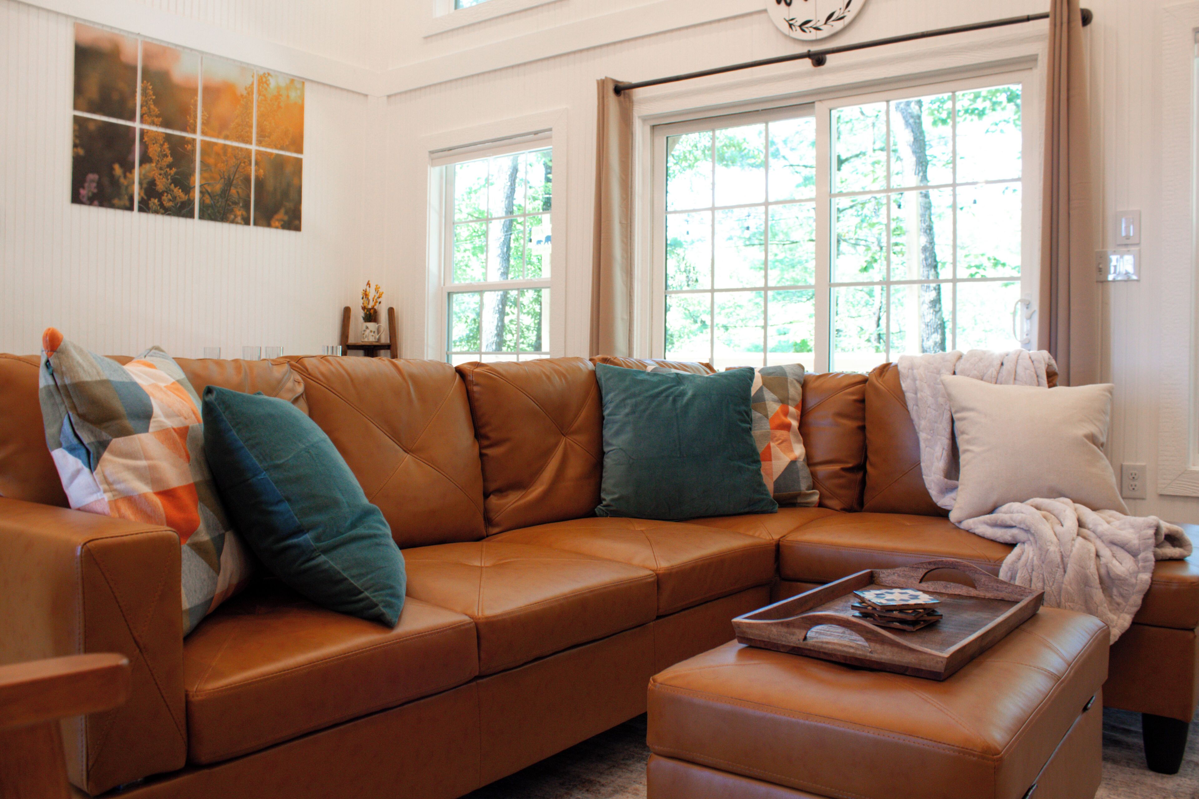 Ruskea nahkainen kulmasohva, jossa on turkoosinvärisiä ja valkoisia koristetyynyjä, puinen tarjotinalusta, jossa on kirja, ja valkoinen peitto samanvärisellä jalkarahilla.