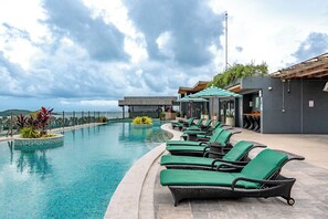A pool area with lounge chairs, a small island with plants, and a building with a modern design. - C214 Calypso unit 102 (Rawai)