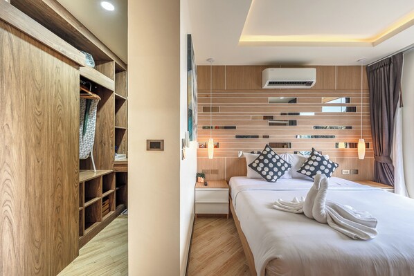 A hotel room with a wooden wardrobe, a neatly made bed with towels, a bedside table, and a wall-mounted air conditioner. - C214 Calypso unit 102 (Rawai)