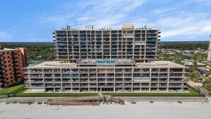 Ein mehrstöckiges Hotel mit Schwimmbad im Erdgeschoss, umgeben von Sandstrand und Grünpflanzen.